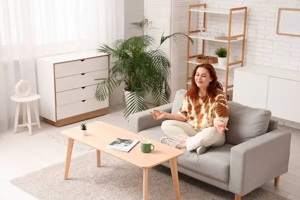 Evdeki kanepede meditasyon yapan genç bir kadın.