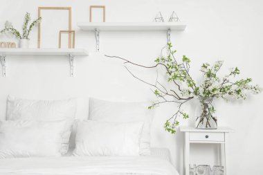 Beyaz yatak odasındaki rahat yatağın yanında, komodinin üzerinde çiçek açan dalları olan vazo.