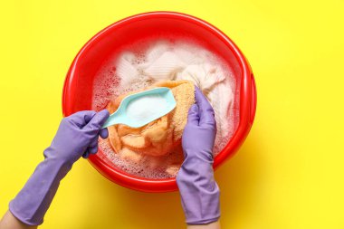 Lastik eldivenli kadın sarı arka plandaki plastik leğende çamaşırlara deterjan ekliyor.