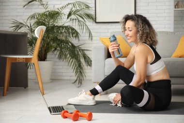 Happy mature sporty woman with laptop, dumbbells and bottle of water at home