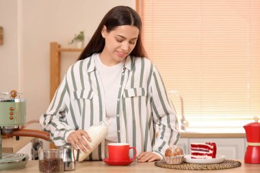 Güzel, genç, mutlu bir kadın mutfakta bir fincan taze kahveye süt dolduruyor.