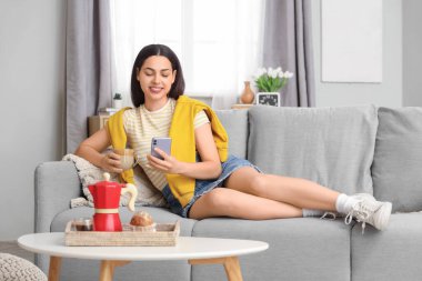 Evinde bir fincan sıcak kahve ve akıllı telefonla güzel genç bir bayan.