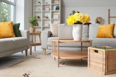 Oturma odasının içinde gri kanepeler, dizüstü bilgisayarlar ve ahşap sehpada sarı narsisli çiçekler.