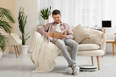 Modern robot vacuum cleaner near sofa with resting young man at home