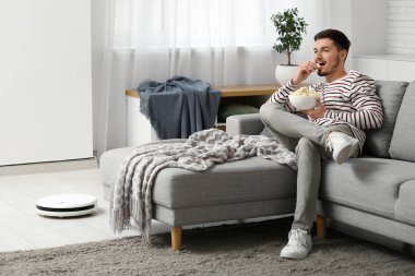 Young man watching movie with modern robot vacuum cleaner at home