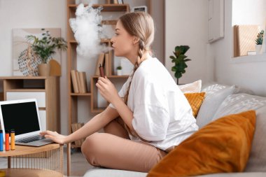 Tek kullanımlık elektronik sigara içen ve evdeki koltukta dizüstü bilgisayar kullanan genç bir kadın.