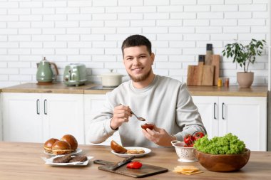 Genç adam mutfakta lezzetli bir burger için ekmeğe ketçap koyuyor.