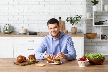 Genç adam mutfakta lezzetli hamburgerin üstüne peynir koyuyor.