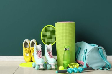 Farklı spor aletleri, patenler ve mavi duvarın yanındaki döşemede spor ayakkabıları.
