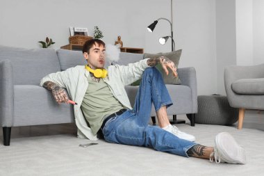 Genç adam tek kullanımlık elektronik sigara içiyor ve evdeki kanepenin yanında oturuyor.