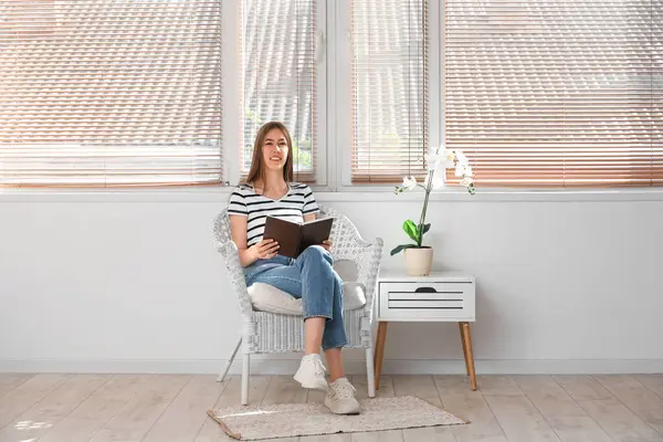 Orkide çiçeğinin yanındaki koltukta oturmuş kitap okuyan genç bir kadın.