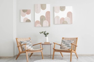 Rahat koltuklar, kahve masası, okaliptüs dallı vazo ve oturma odasındaki beyaz duvarda güzel resimler.