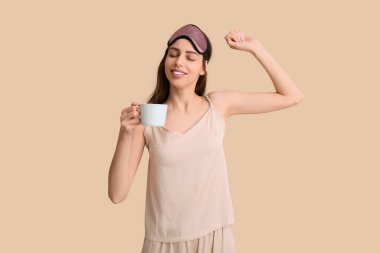 Uyku maskesi ve bej arka planda bir fincan kahve olan pijamalı genç bir kadın.