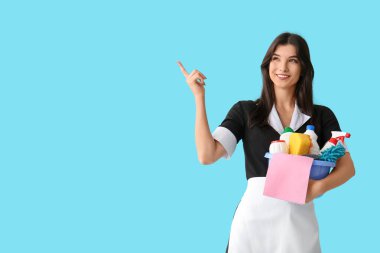 Temizlik malzemeleriyle genç oda hizmetçisi mavi arka plandaki bir şeyi işaret ediyor.