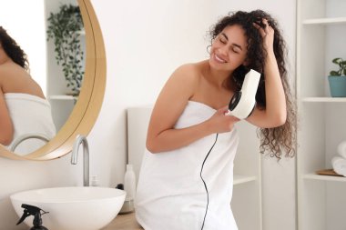 Güzel, genç, mutlu, Afro-Amerikan bir kadın. Sağlıklı kıvırcık saçlarını banyoda kurutuyor.