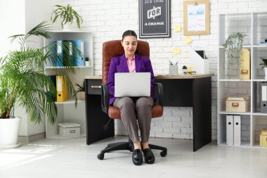 Dizüstü bilgisayar kullanan ve işyerinin yanındaki koltukta oturan kadın ofis çalışanı.