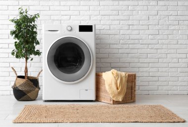 Beyaz tuğla duvarın yanında ev bitkisi, sepet ve çamaşırları olan modern çamaşır makinesi.