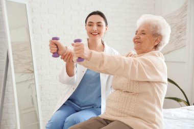 Fizyoterapist ve yaşlı kadın yatak odasında dambıllarla antrenman yapıyor.