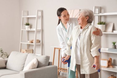 Koltuk değnekleriyle ve fizyoterapistiyle evde yürüyen yaşlı bir kadın.