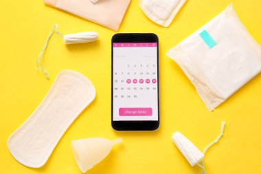 Cep telefonu ekranında adet takvimi ve sarı arka planda kadın hijyen ürünleri