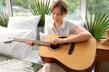 Genç bir kadın yatak odasında gitar çalıyor.