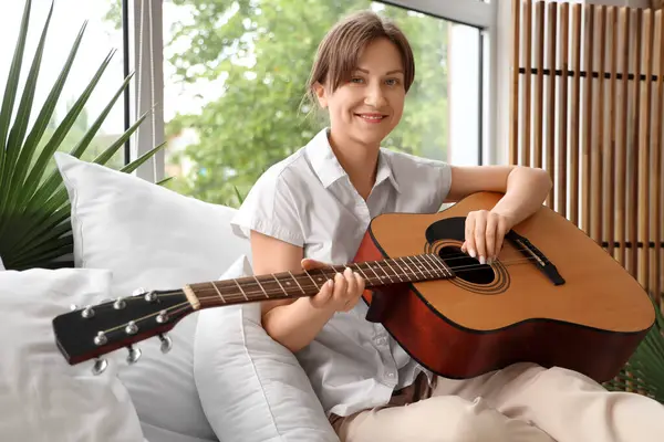 Genç bir kadın yatak odasında gitar çalıyor.