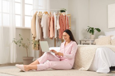 Yatak odasındaki elbise rafının yanında dizüstü bilgisayar kullanan genç bir kadın.