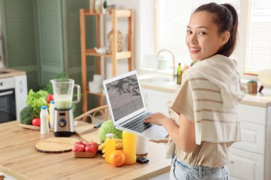 Genç Asyalı kadın mutfakta dizüstü bilgisayar kullanıyor.