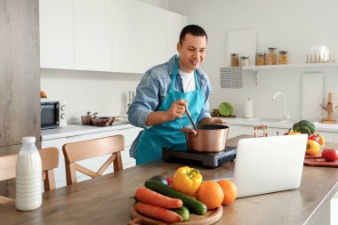 Dizüstü bilgisayarlı genç adam mutfakta çorba pişiriyor.