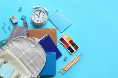 Farklı kırtasiye malzemelerinin olduğu okul çantası ve mavi arka planda çalar saat.