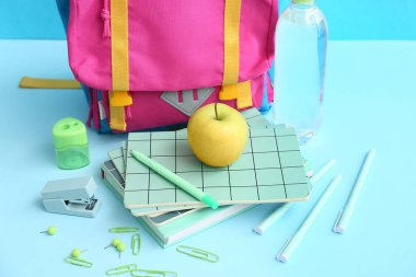 Farklı kırtasiye malzemeleriyle renkli okul çantası, mavi arka planda bir şişe su ve elma.