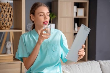 Genç bir tıp öğrencisi, elinde dosya ve bir bardak suyla evdeki kanepede oturuyor.