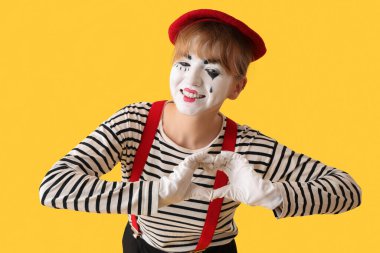 Kadın pandomimci elleri sarı arka planda kalp yapıyor.