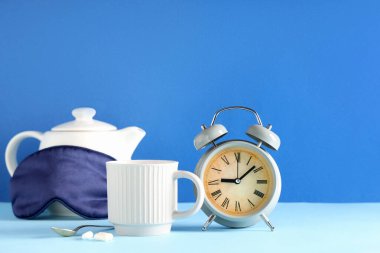 Bir fincan çay, çalar saat, uyku maskesi ve mavi duvarın yanındaki masada haplar.