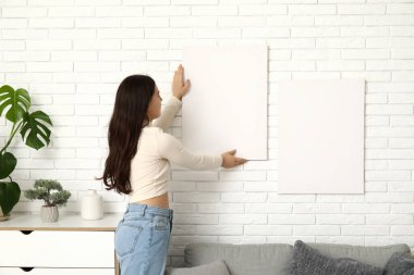 Oturma odasındaki beyaz tuğla duvara boş tuval asan genç bir kadın.