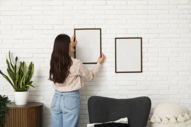 Genç bir kadın oturma odasındaki beyaz tuğla duvara boş bir çerçeve asıyor. Arka plan