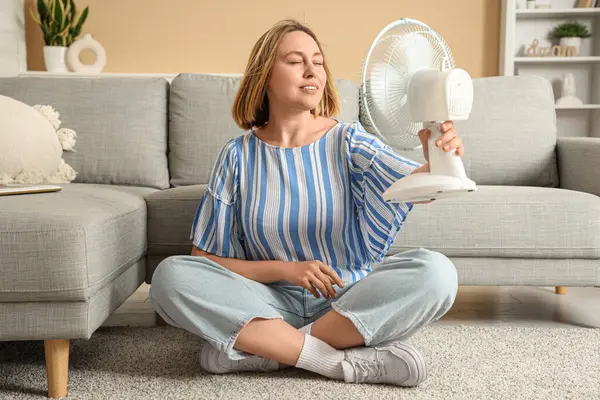 Güzel, olgun, mutlu bir kadın elektrikli vantilatörüyle evde oturuyor.