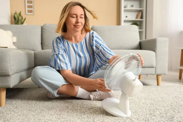 Güzel, olgun, mutlu bir kadın elektrikli vantilatörüyle evde oturuyor.