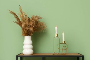 Oturma odasındaki yeşil duvarın yanındaki şifonyerin üzerinde vazo dolusu otla mum yakmak.