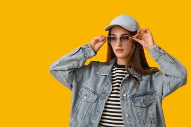 Şık güneş gözlüklü ve sarı arka planda şapkalı güzel bir genç kadın.
