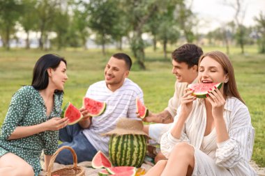 Genç bir kadın arkadaşlarıyla parkta piknik yaparken karpuz yiyor.
