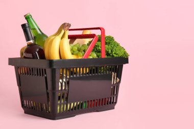 Farklı taze ürünlerle dolu siyah alışveriş sepeti ve pembe arka planda muzlar.