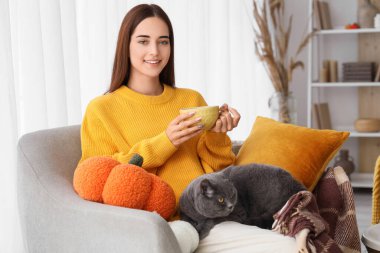 Bir fincan yeşil çay ve tatlı İngiliz kedisiyle sonbahar günü evinde koltuğunda oturan genç bir kadın.