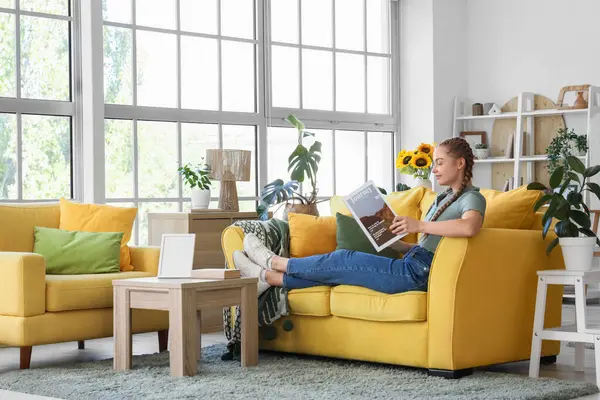 Evdeki sarı kanepede dergi okuyan genç bir kadın.