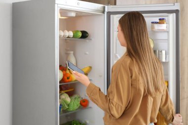 Cep telefonu olan genç bir kadın mutfaktaki açık buzdolabının yanında.