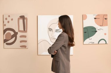 Sanat galerisindeki bej duvardaki resimlere bakan genç bir kadın.