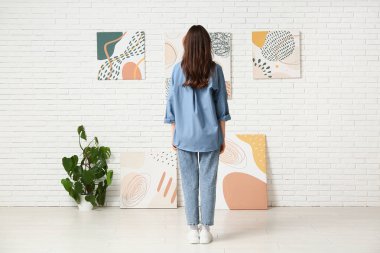Sanat galerisinde beyaz tuğlalı duvardaki resimlere bakan genç bir kadın.