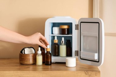 Kadın eli, bej arka planda küçük kozmetik buzdolabı olan masadan serum alıyor. Yakın çekim.