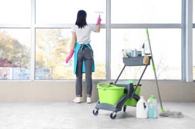 Odada temizlik malzemeleriyle dolu bir tramvayla pencereyi temizleyen kadın hademe.