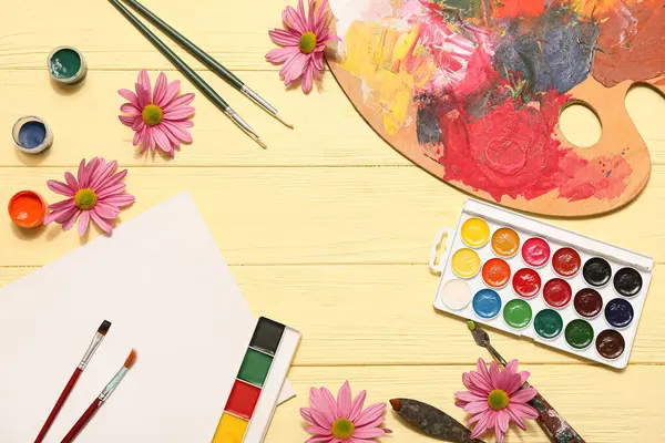 Sanatçı fırçalarından, paletten, boyalardan ve sarı ahşap arka plandaki çiçeklerden yapılmış çerçeve